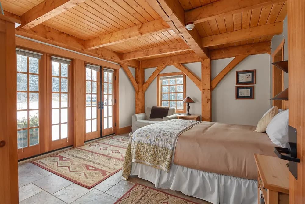 Cozy bedroom with wooden beams, a bed, and large windows leading to an outdoor view.