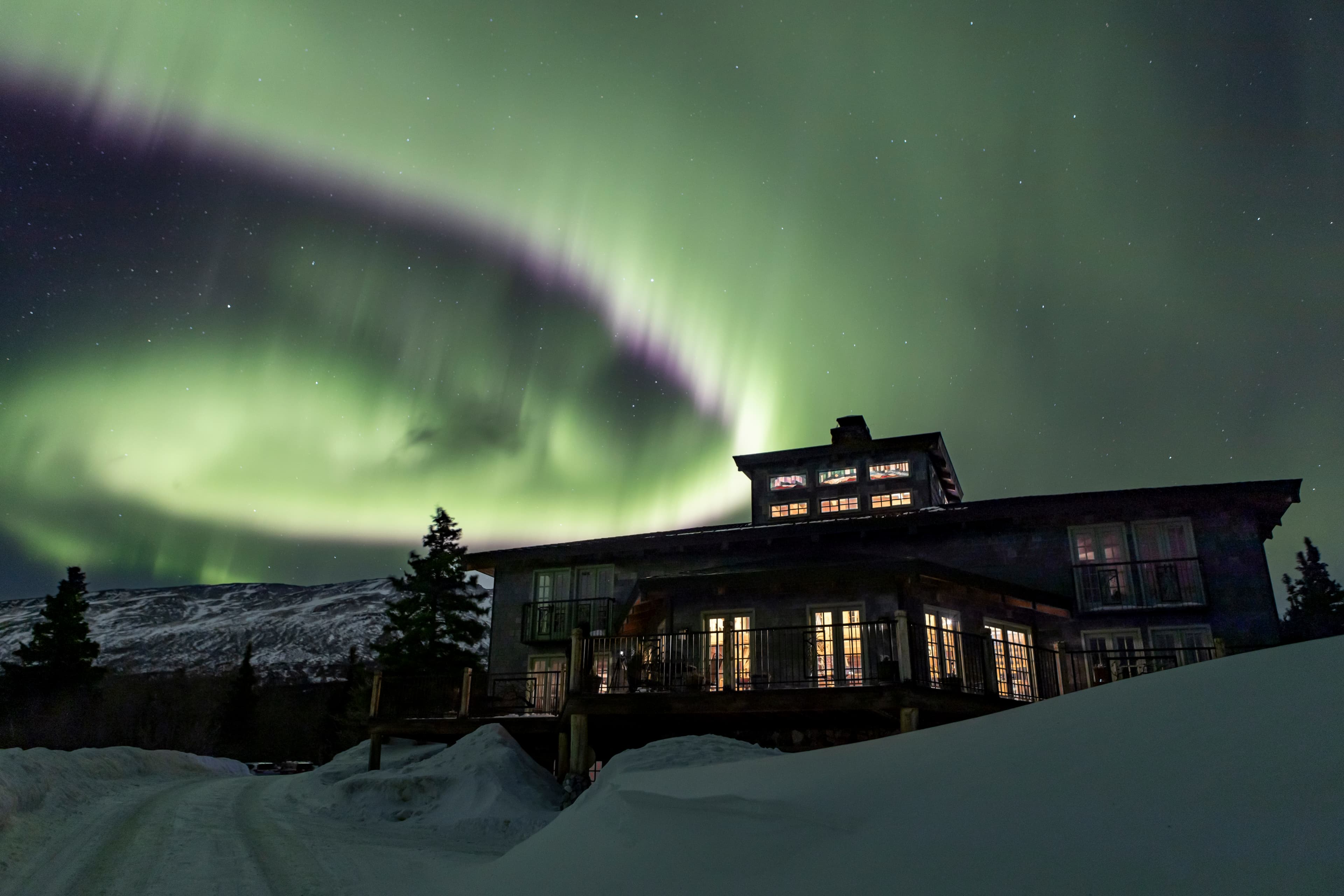 A cozy lodge sits beneath a vibrant display of the northern lights in a snowy landscape.