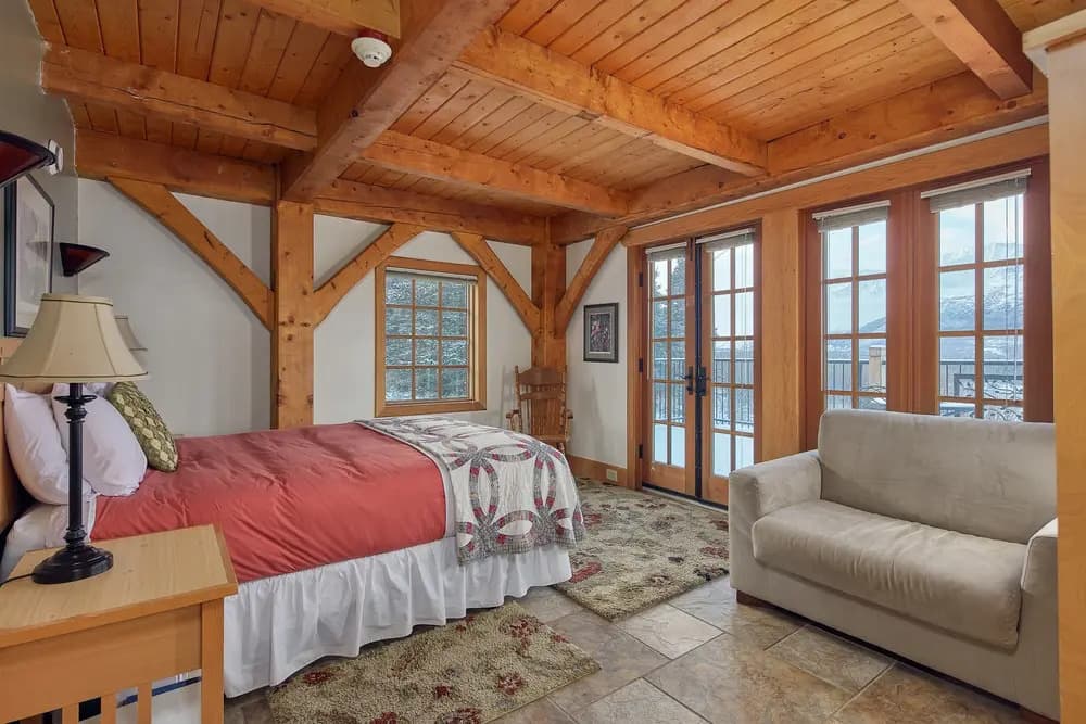 A cozy bedroom with wooden beams, a large bed, a sofa, and windows overlooking a snowy landscape.