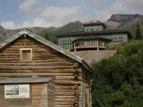 A rustic wooden cabin in the foreground with a modern building and mountains in the background.