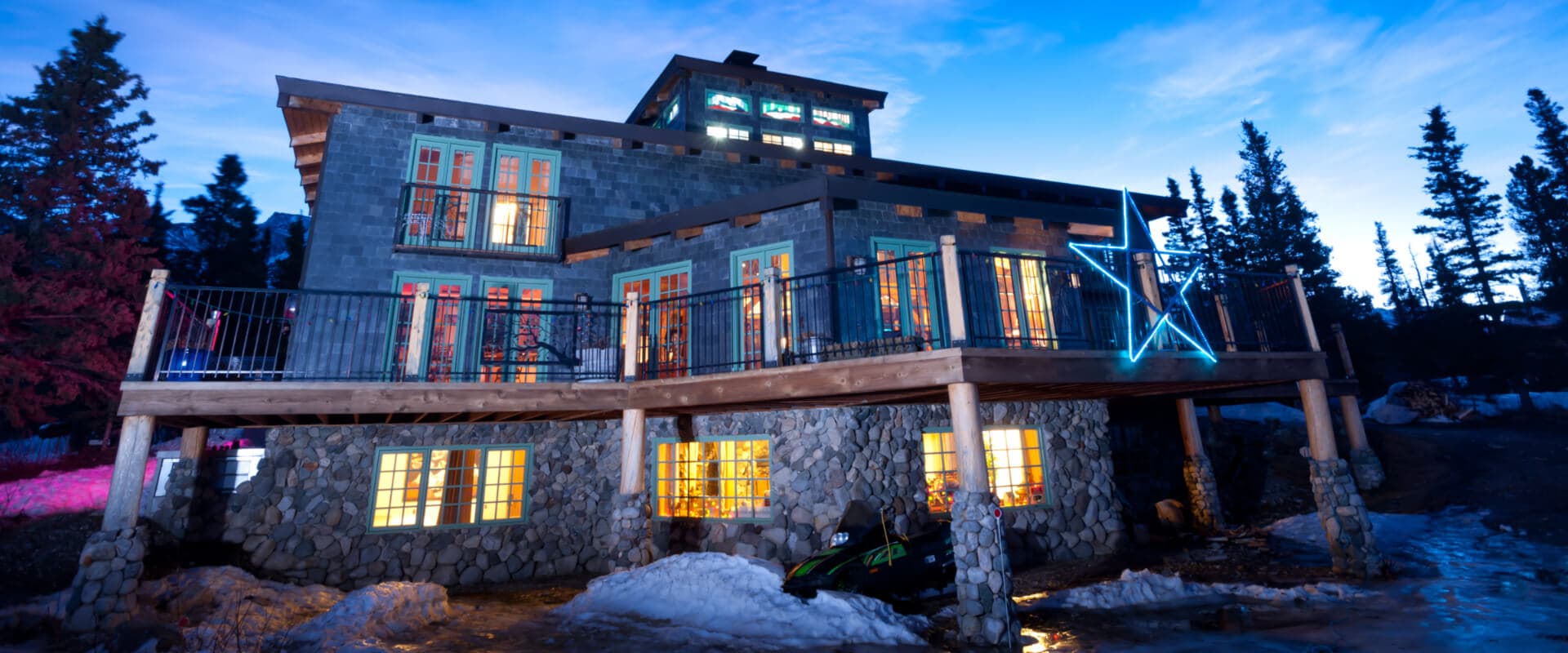 A modern stone house with large windows and a balcony, illuminated against a twilight sky.