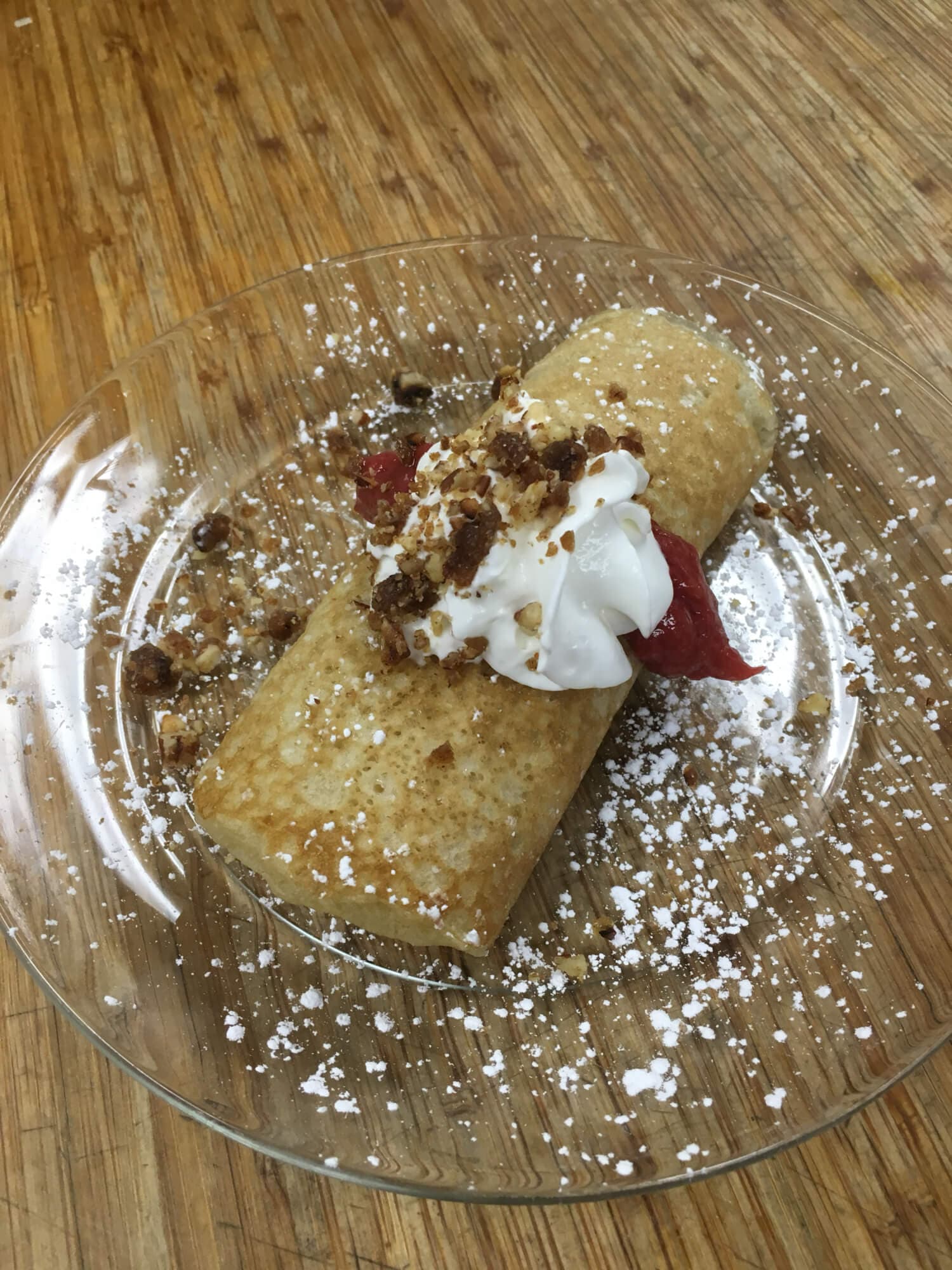 A golden dessert crepe topped with whipped cream, syrup, and crumbled toppings, served on a glass plate.