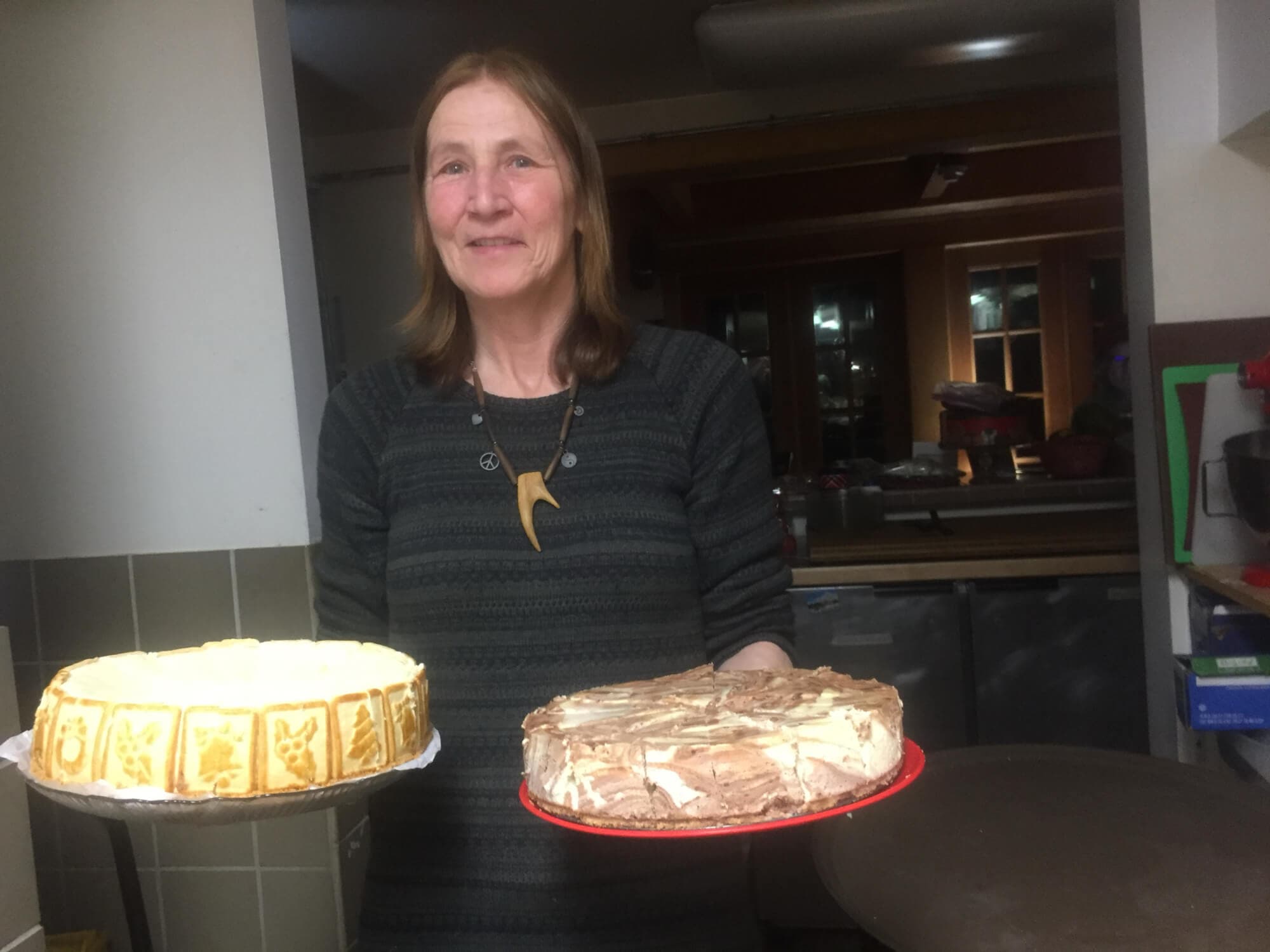 A woman smiles while holding two beautifully decorated cakes in a kitchen.
