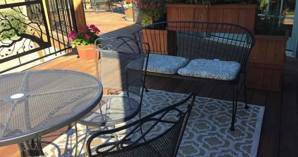 A cozy outdoor seating area with a circular table, metal chairs, and a cushioned bench amidst potted flowers.