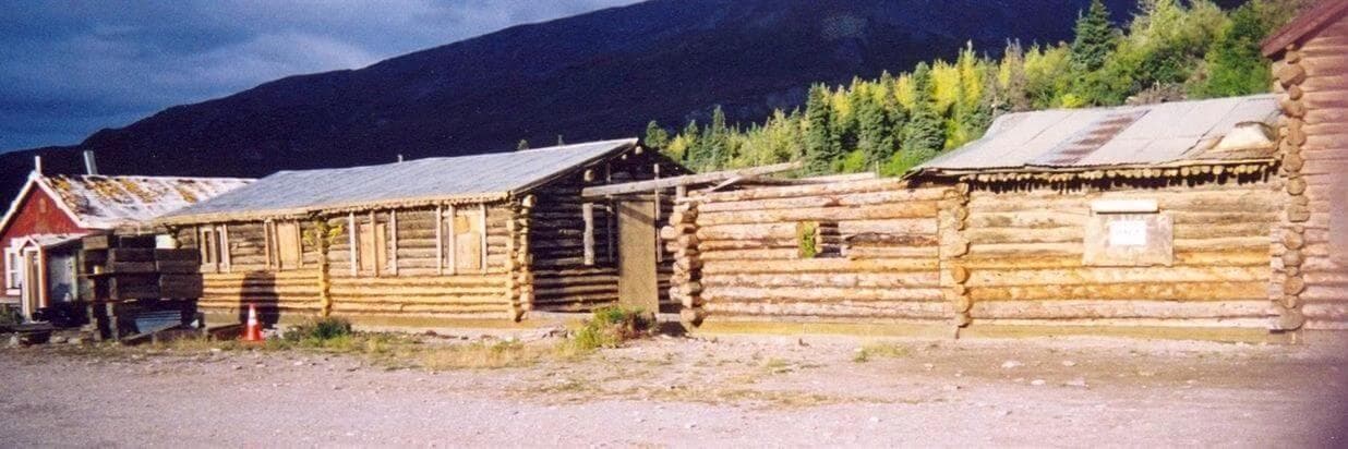 A row of rustic log cabins surrounded by trees and mountains.