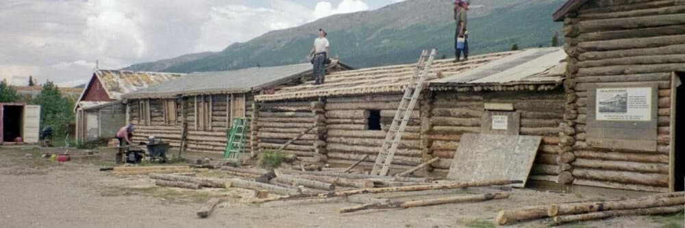 Men working on the roof of a log building in a rural setting.