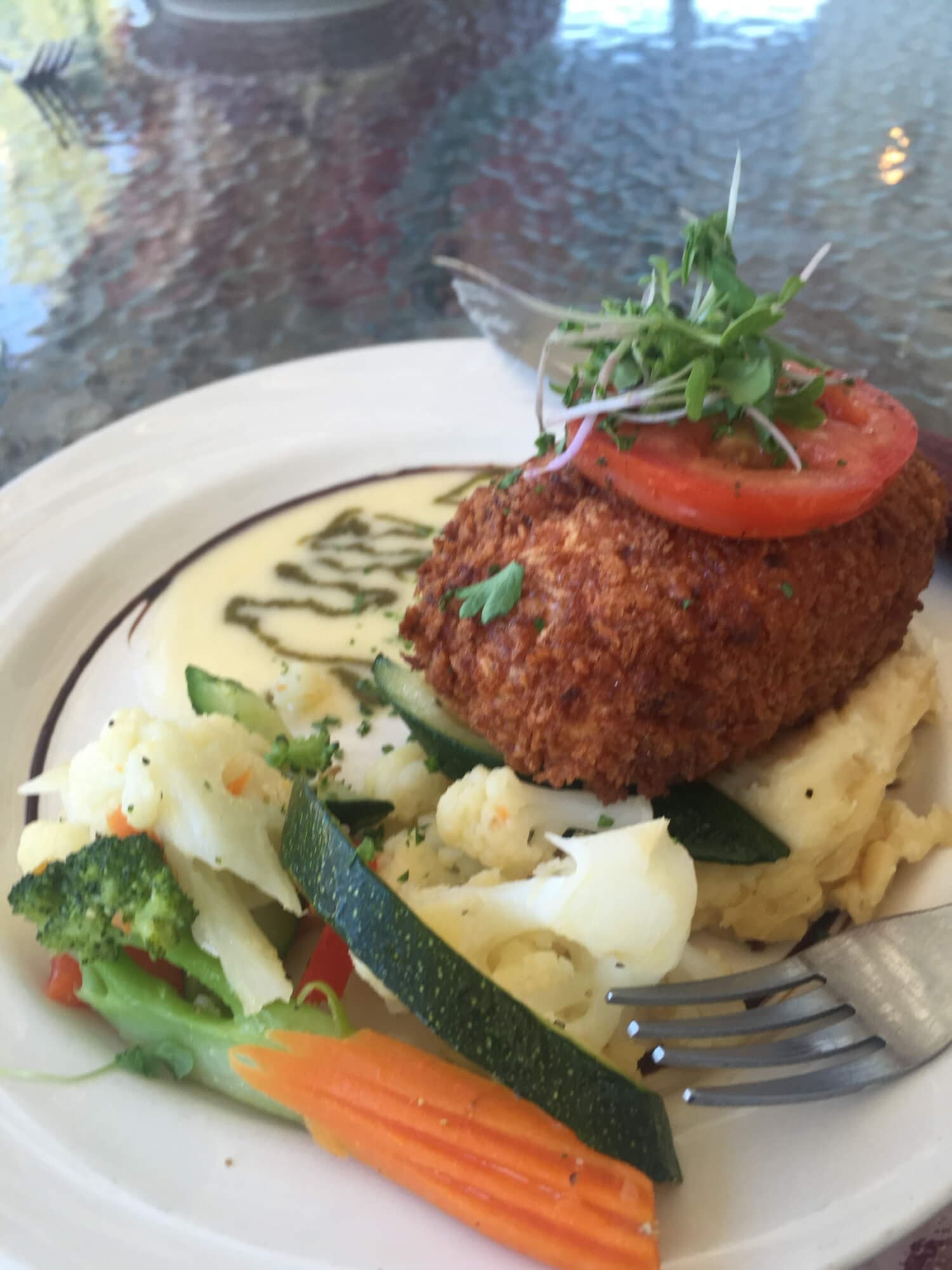 A plate of crispy fried fish topped with a tomato slice and microgreens, served with mashed potatoes and mixed vegetables.