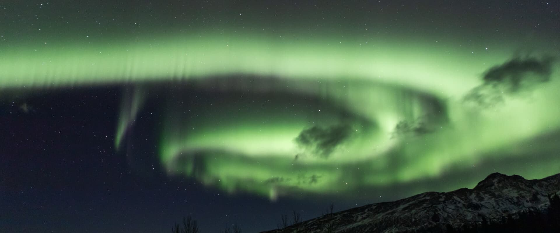 A swirling display of green auroras lights up the night sky above a silhouette of mountains.