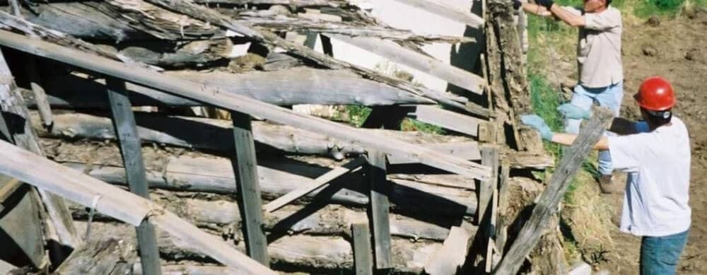 Two individuals work on a dilapidated wooden structure, one holding a beam while the other inspects the repairs.