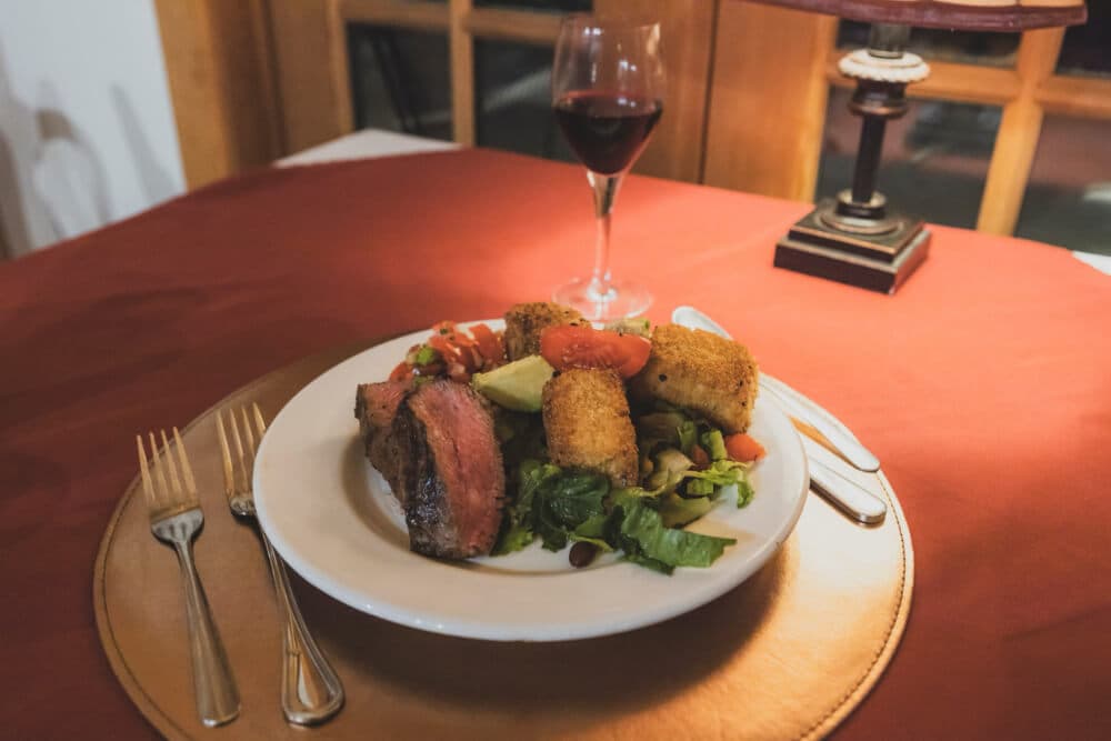 A plate of steak, salad, and croquettes accompanied by a glass of red wine on a table with a red tablecloth.
