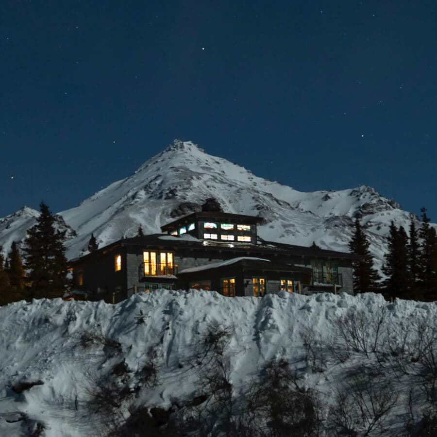 A snow-covered mountain house illuminated at night, surrounded by trees and peaks under a starry sky.