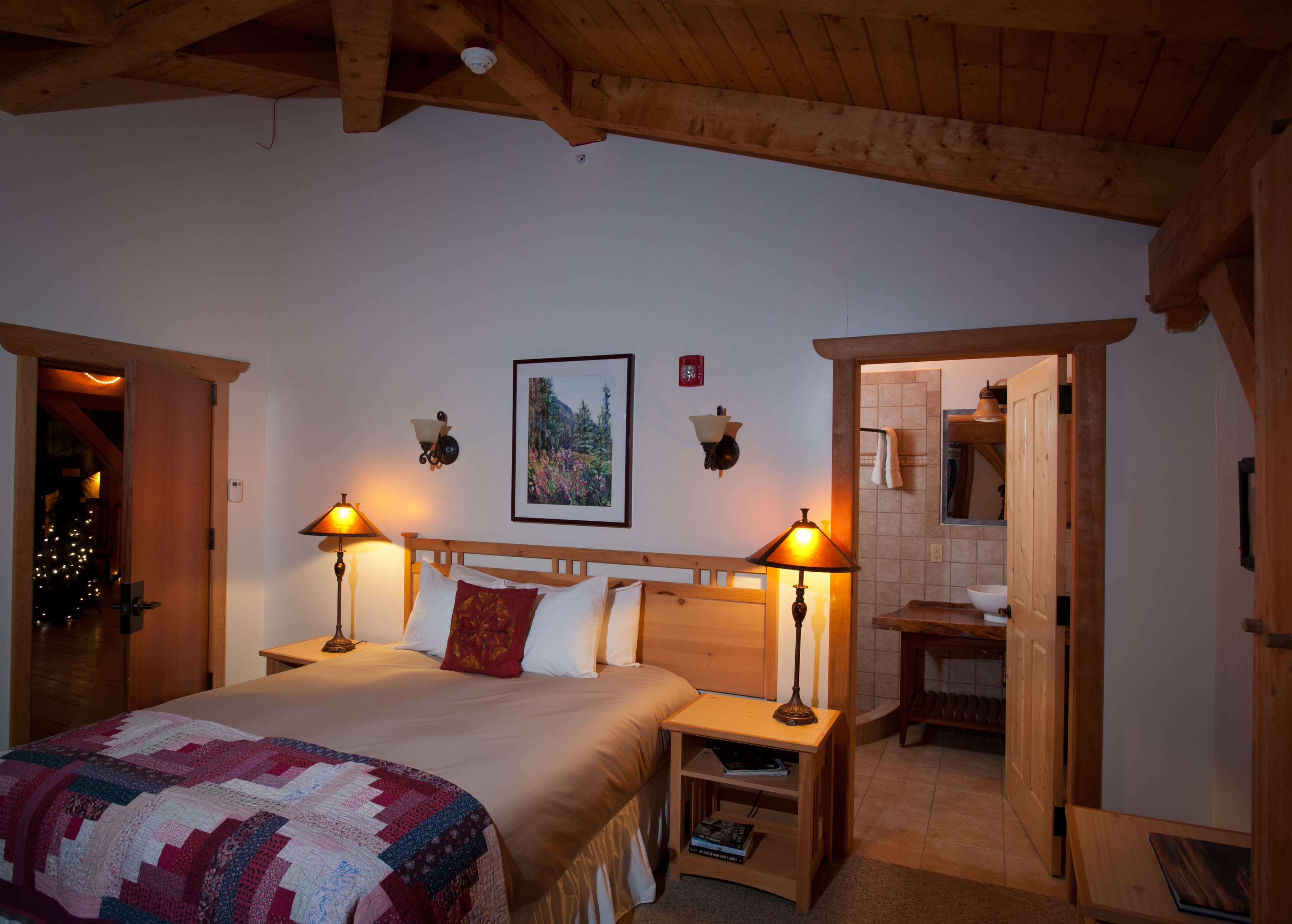 A cozy bedroom with a wooden ceiling, featuring a bed with a quilt, two bedside lamps, and an adjoining bathroom.