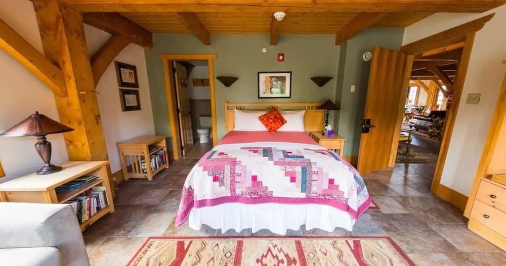 A cozy bedroom featuring a bed with a colorful quilt, wooden beams, and a warm ambiance.
