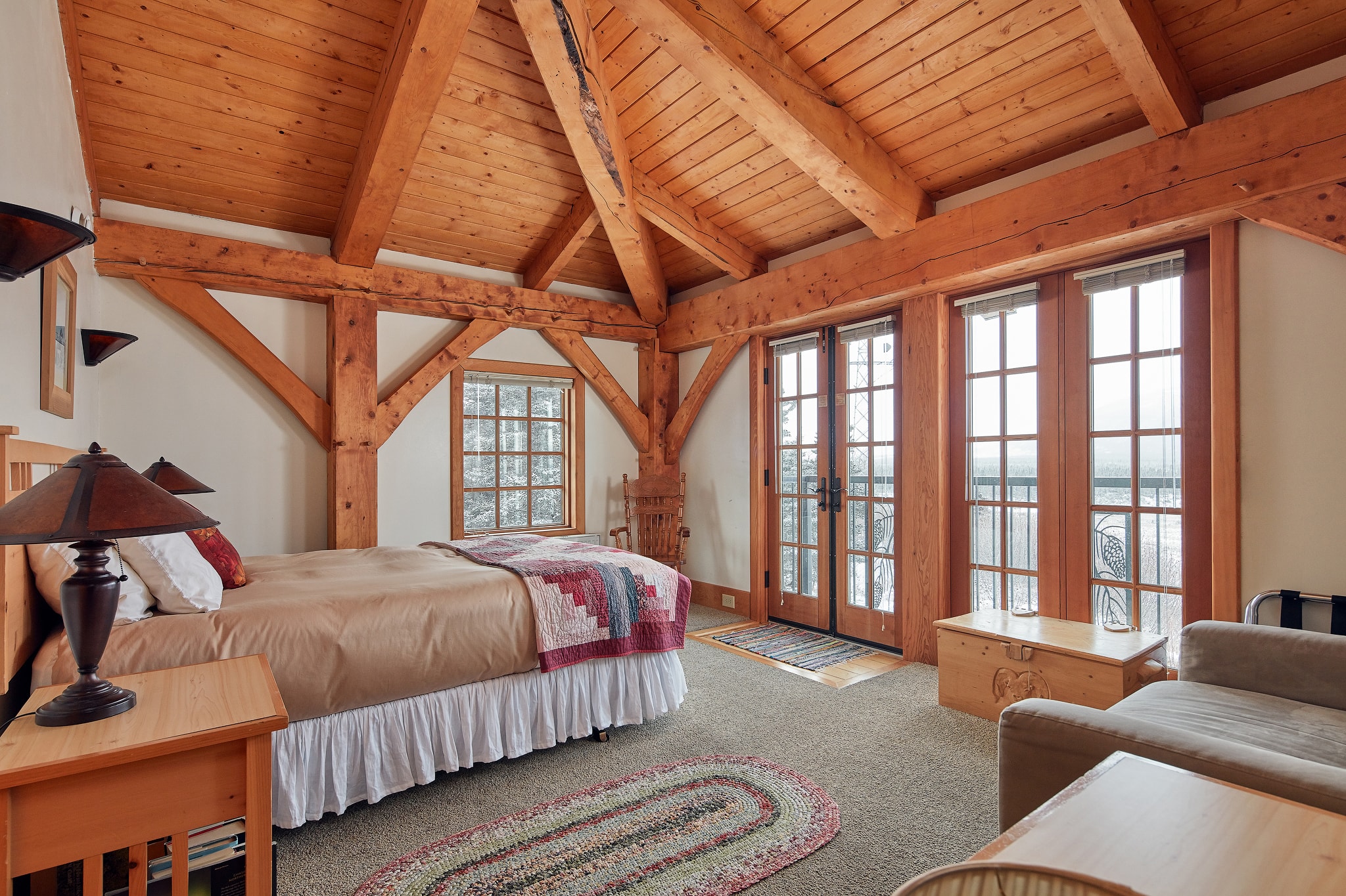 Cozy bedroom featuring wooden beams, a queen bed, and large windows with a small seating area.