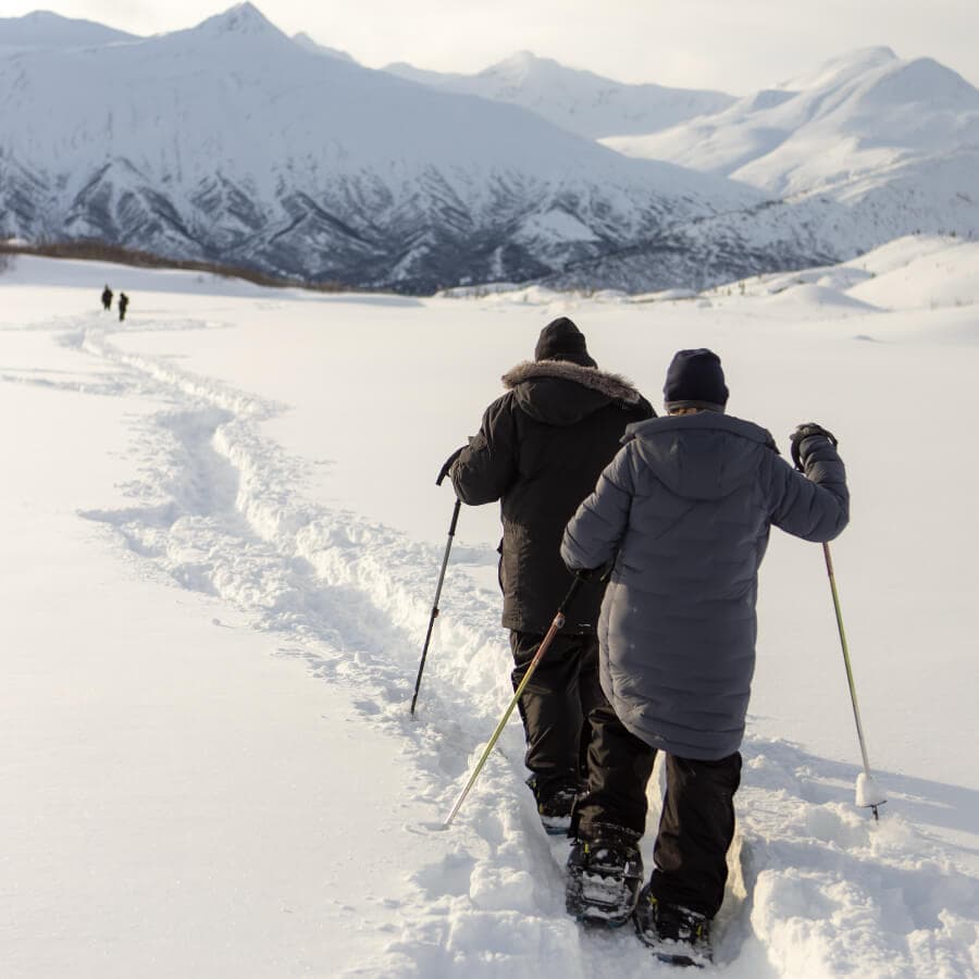 Two people trek through snowy landscape with mountains in the background.