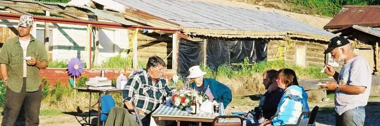 A group of people gather around a table outdoors, enjoying snacks and conversation near rustic buildings.