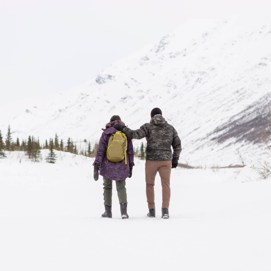 Two people walking together in a snowy landscape, with mountains and trees in the background.