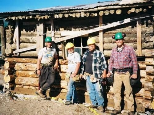 Four construction workers in hard hats stand in front of a log building.