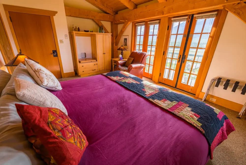 Cozy bedroom with a purple bedspread, colorful pillows, and large windows overlooking a scenic view.