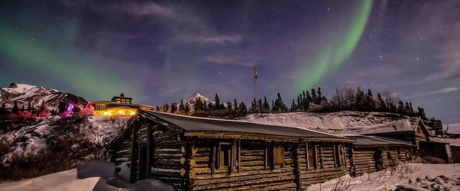 A rustic log cabin sits under a vibrant aurora borealis in a snowy landscape.