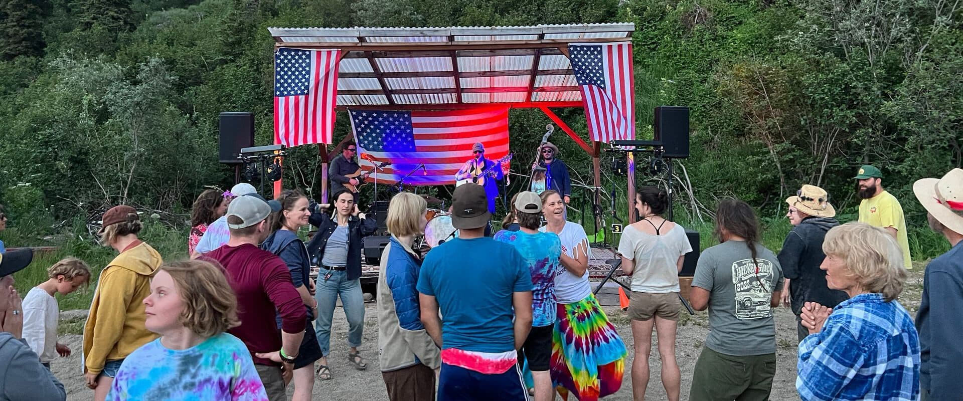 A lively outdoor concert featuring performers on stage with American flags, surrounded by an engaged audience.